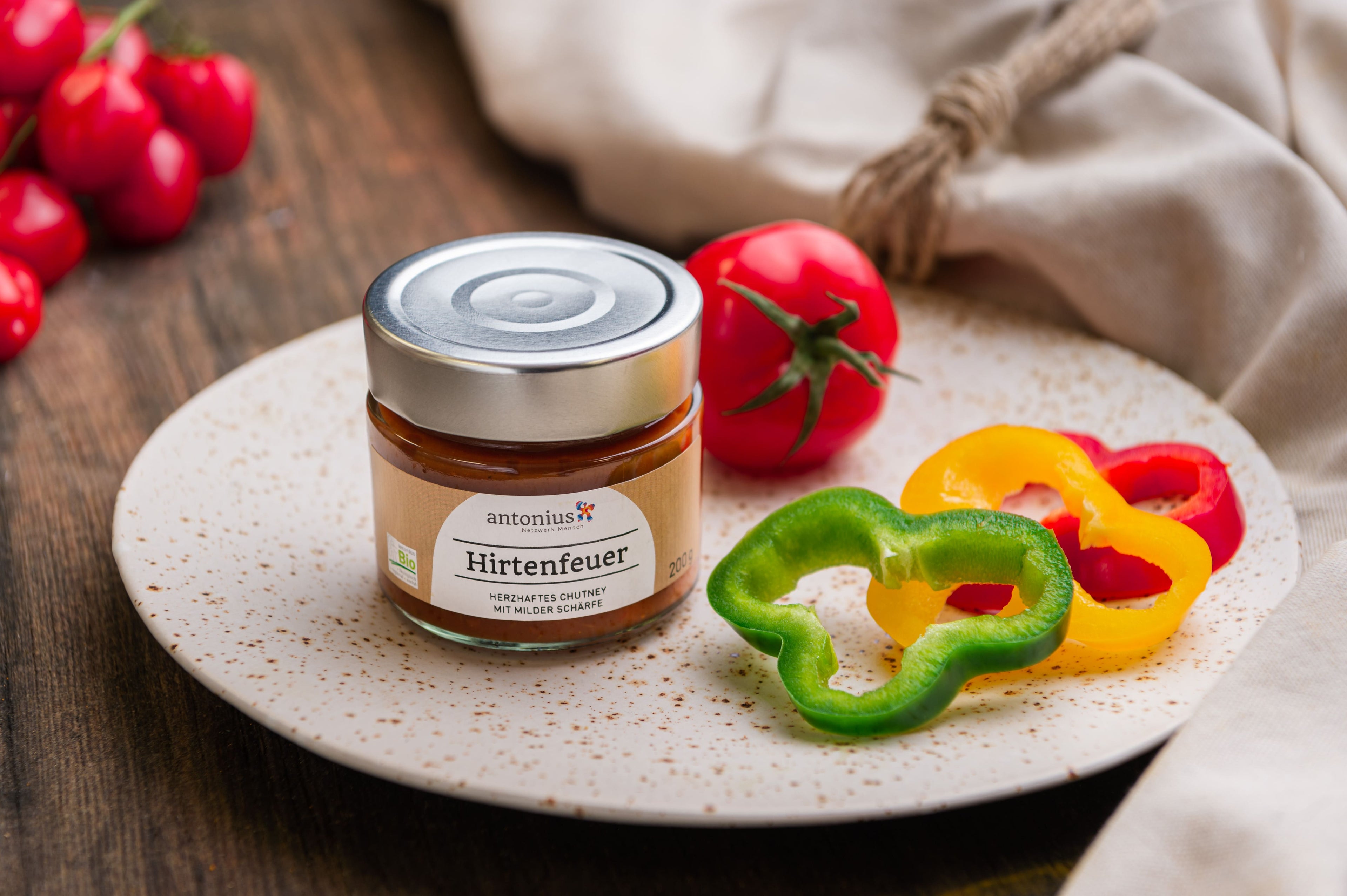 Offenes Glas Hirtenfeuer-Chutney auf einem Teller, dekoriert mit drei verschieden farbigen Paprika und einer Tomate.