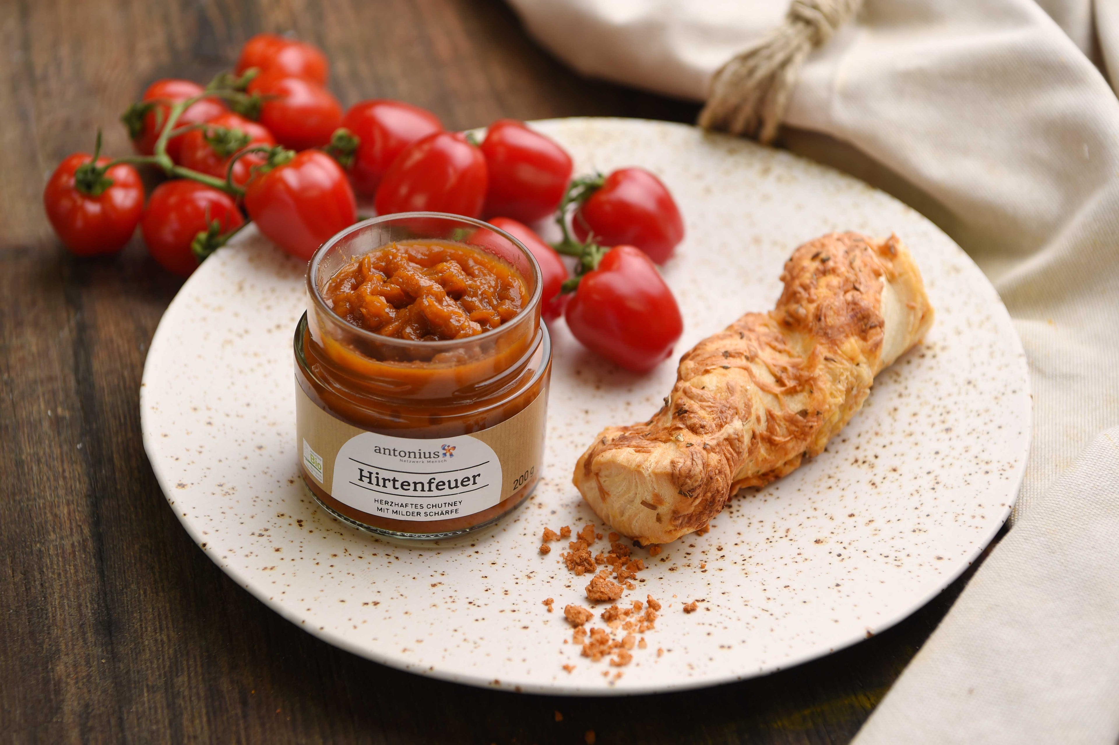 Offenes Glas Hirtenfeuer-Chutney auf einem Teller, dekoriert mit einer Käsestange und Tomaten.