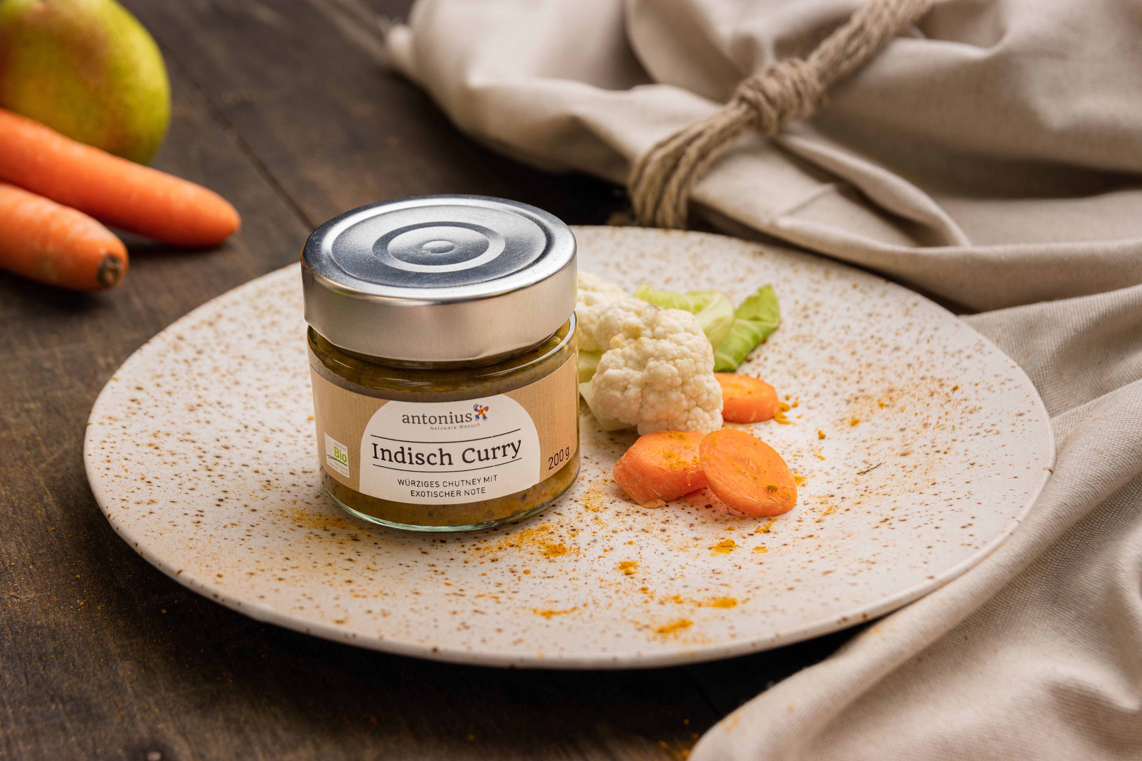 Ein Glas Indisch-Curry Chutney auf einem Teller, dekoriert mit Blumenkohl, Salat und Karotten.