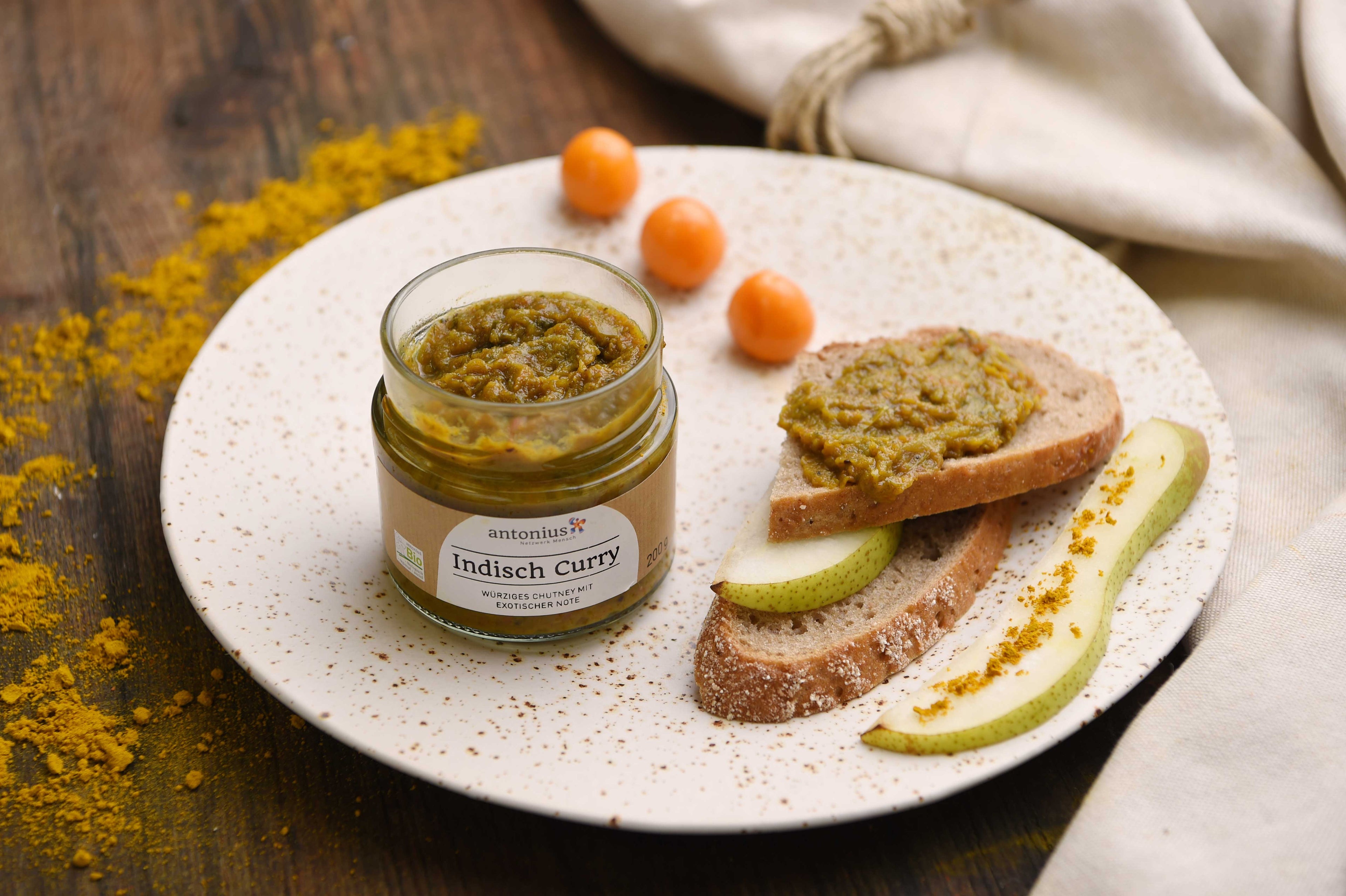 Ein offenes Glas Indisch-Curry Chutney auf einem Teller, dekoriert mit Birne. Physalis und mit dem Chutney bestrichenem Brot.