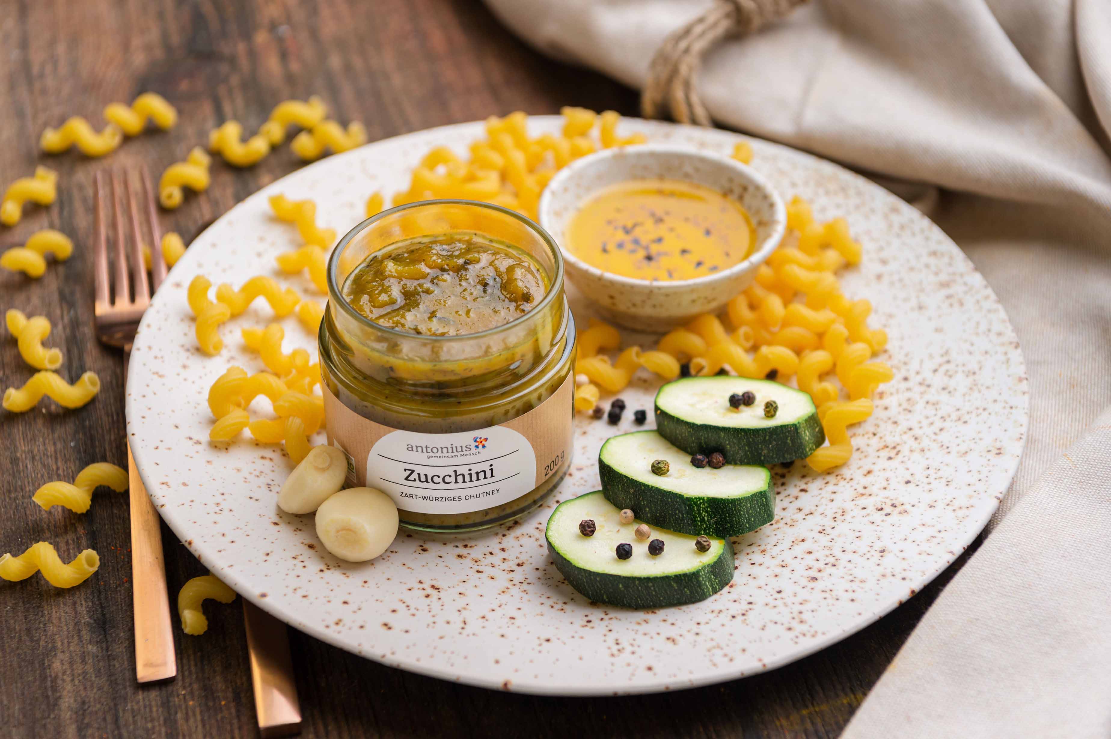Ein offenes Glas Zucchini-Chutney auf einem Teller, angerichtet mit Nudeln, Zucchini, Pfefferkörnern und Knoblauchzehen.