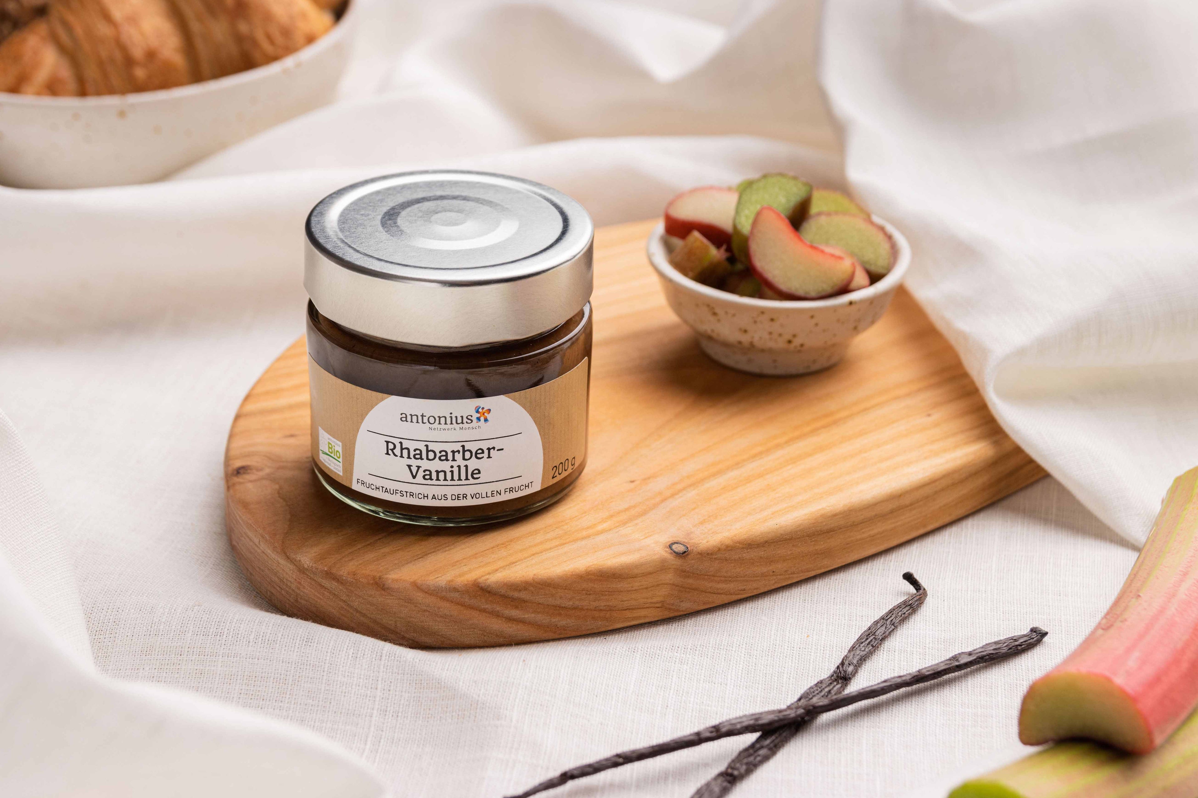 Ein Glas Rhabarber-Vanille-Fruchtaufstrich auf einem Frühstücksbrettchen, angerichtet mit geschnittenem Rhabarber und Vanilleschoten.