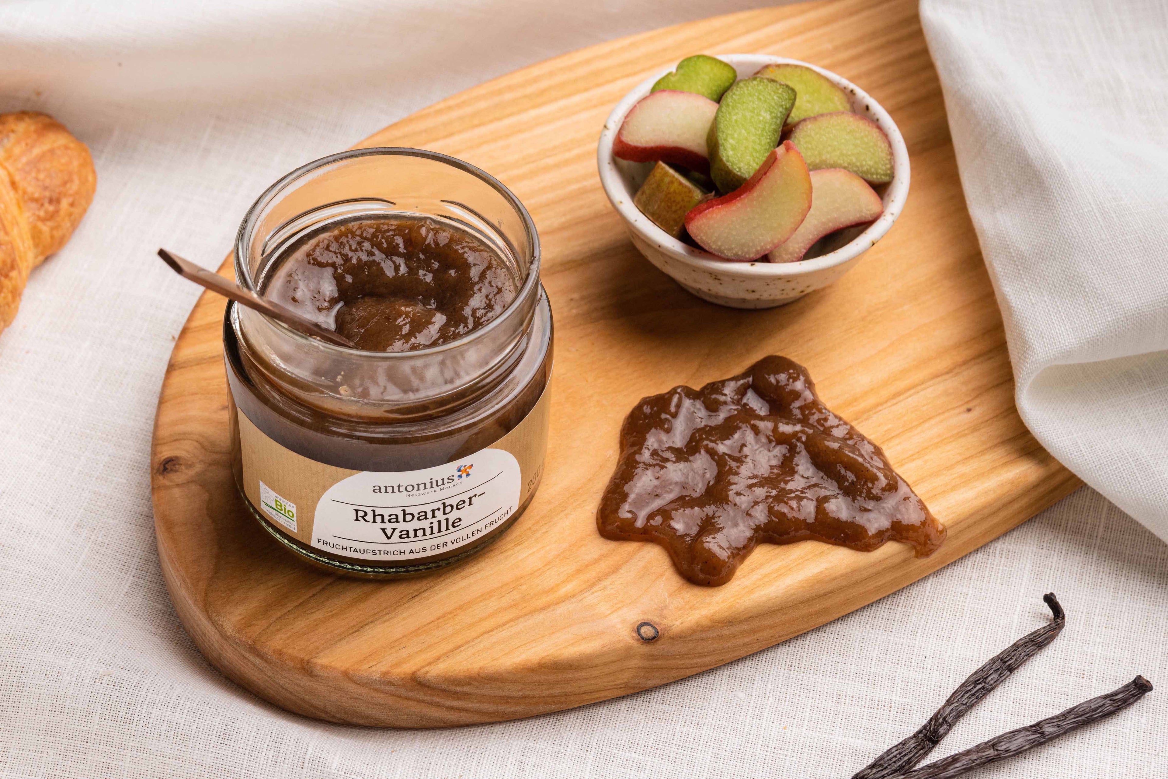 Ein offenes Glas Schwarze Johannisbeere-Fruchtaufstrich auf einem Frühstücksbrettchen, angerichtet mit klein geschnittenem Rhabarber und einem Klecks Fruchtaufstrich.