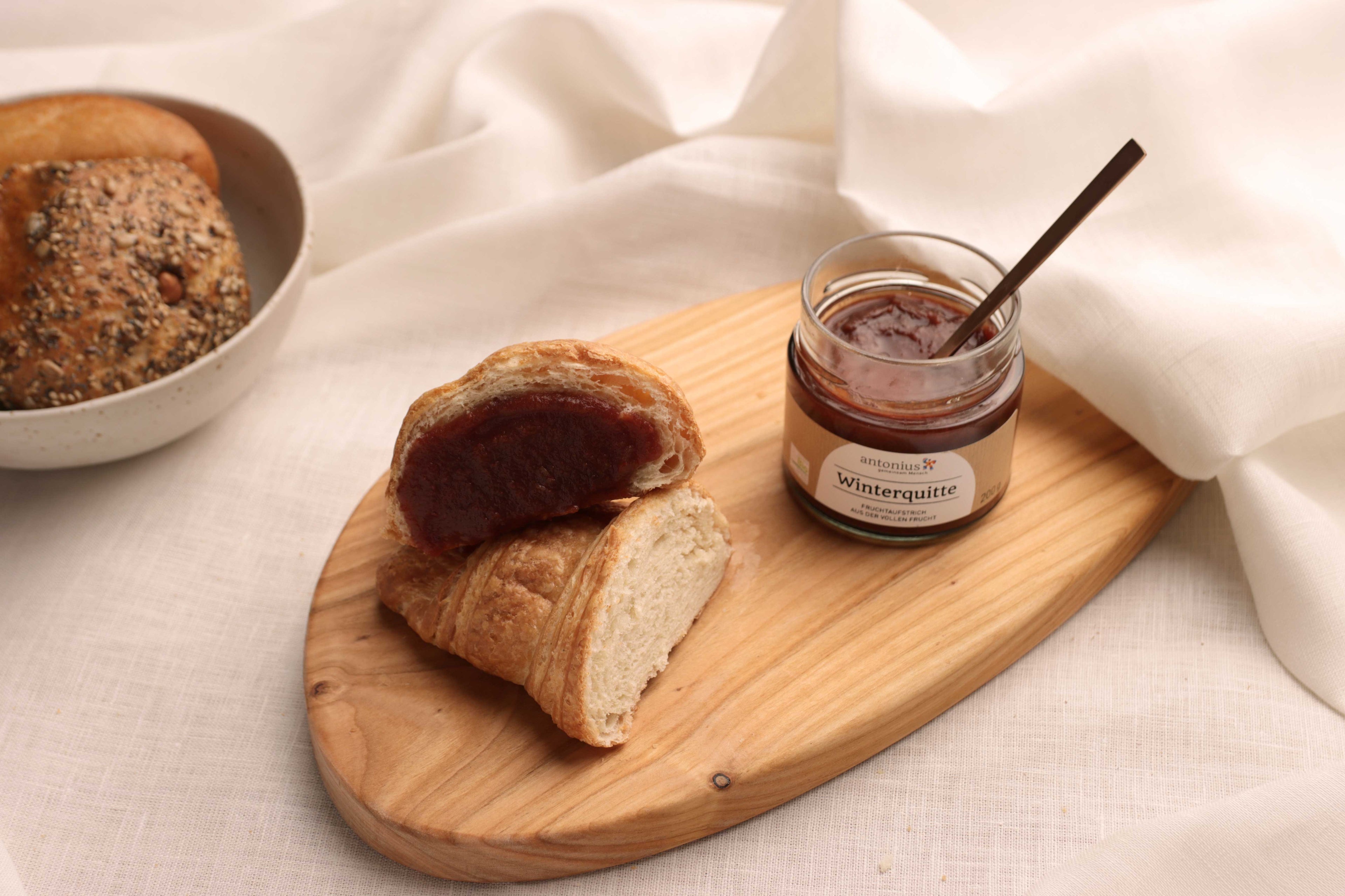 Ein offenes Glas Winterquitten-Fruchtaufstrich auf einem Frühstücksbrettchen, angerichtet neben einem Croissant, das mit Aufstrich bestrichen ist.