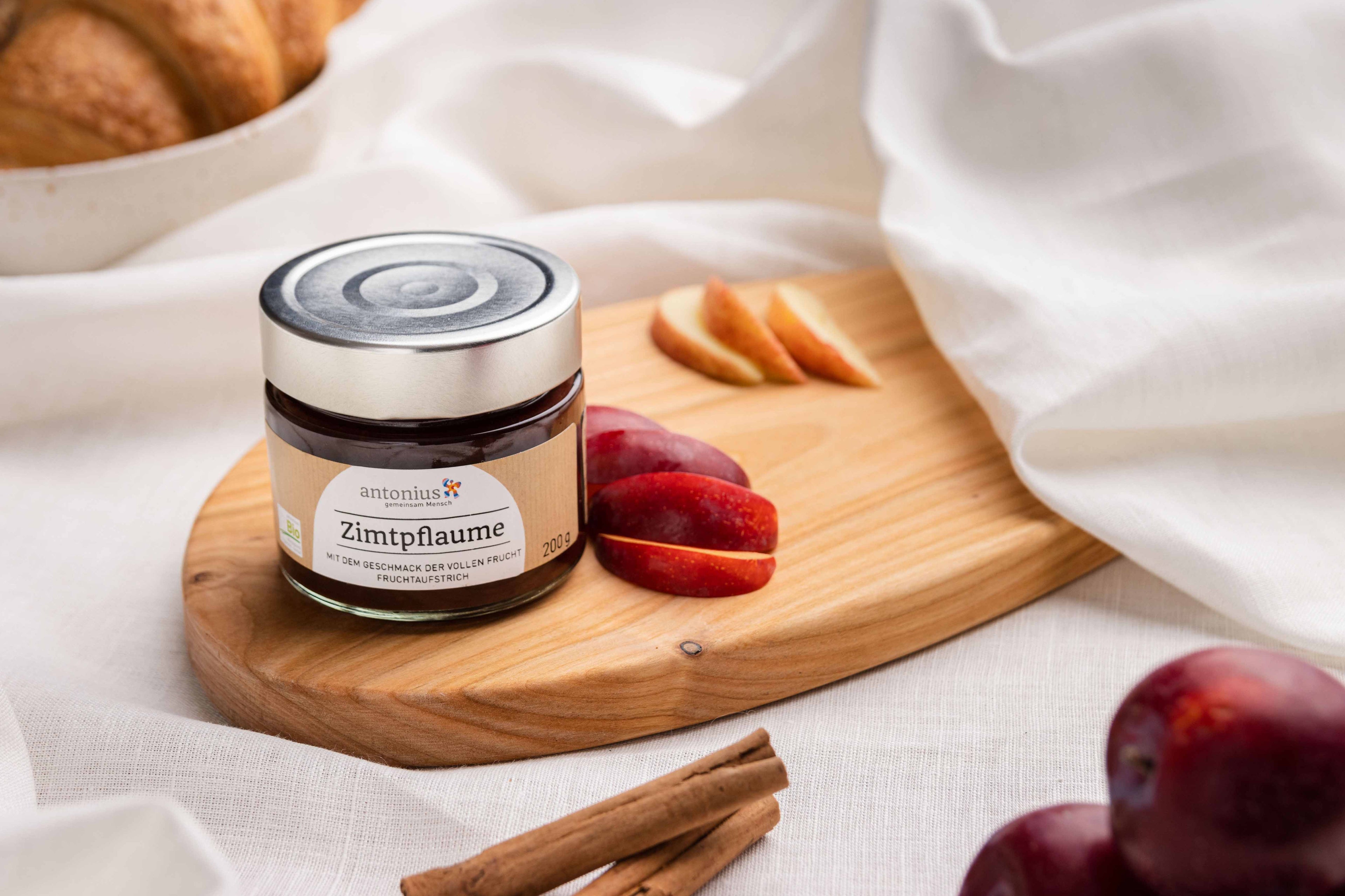 Ein Glas Zimtpflaume-Fruchtaufstrich auf einem Frühstücksbrettchen, angerichtet mit Pflaumenscheiben und Zimt.