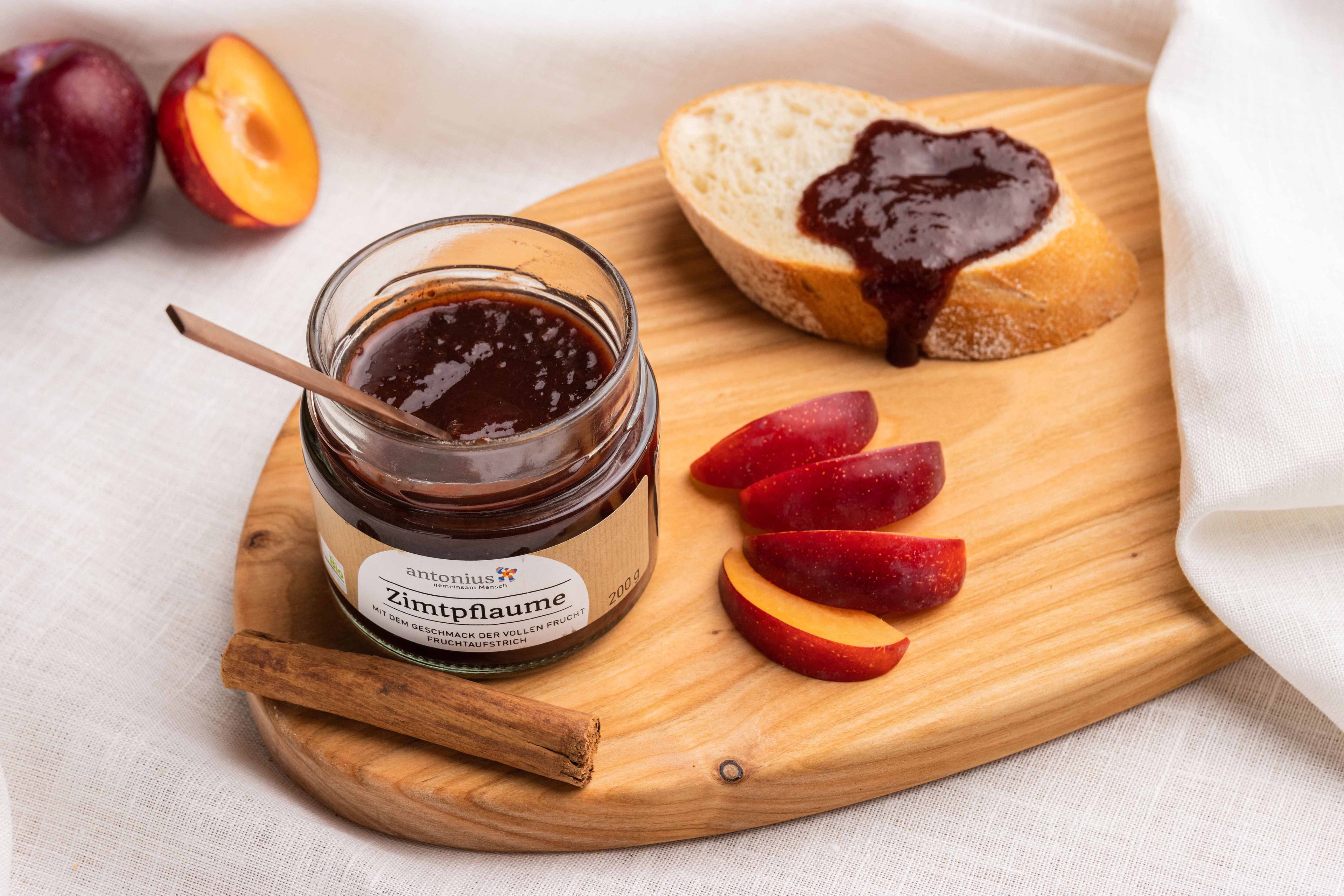 Ein offenes Glas Zimtpflaume-Fruchtaufstrich auf einem Frühstücksbrettchen, angerichtet mit Pflaumenscheiben, Zimt und einer Baguettescheibe, die mit Aufstrich bestrichen ist.