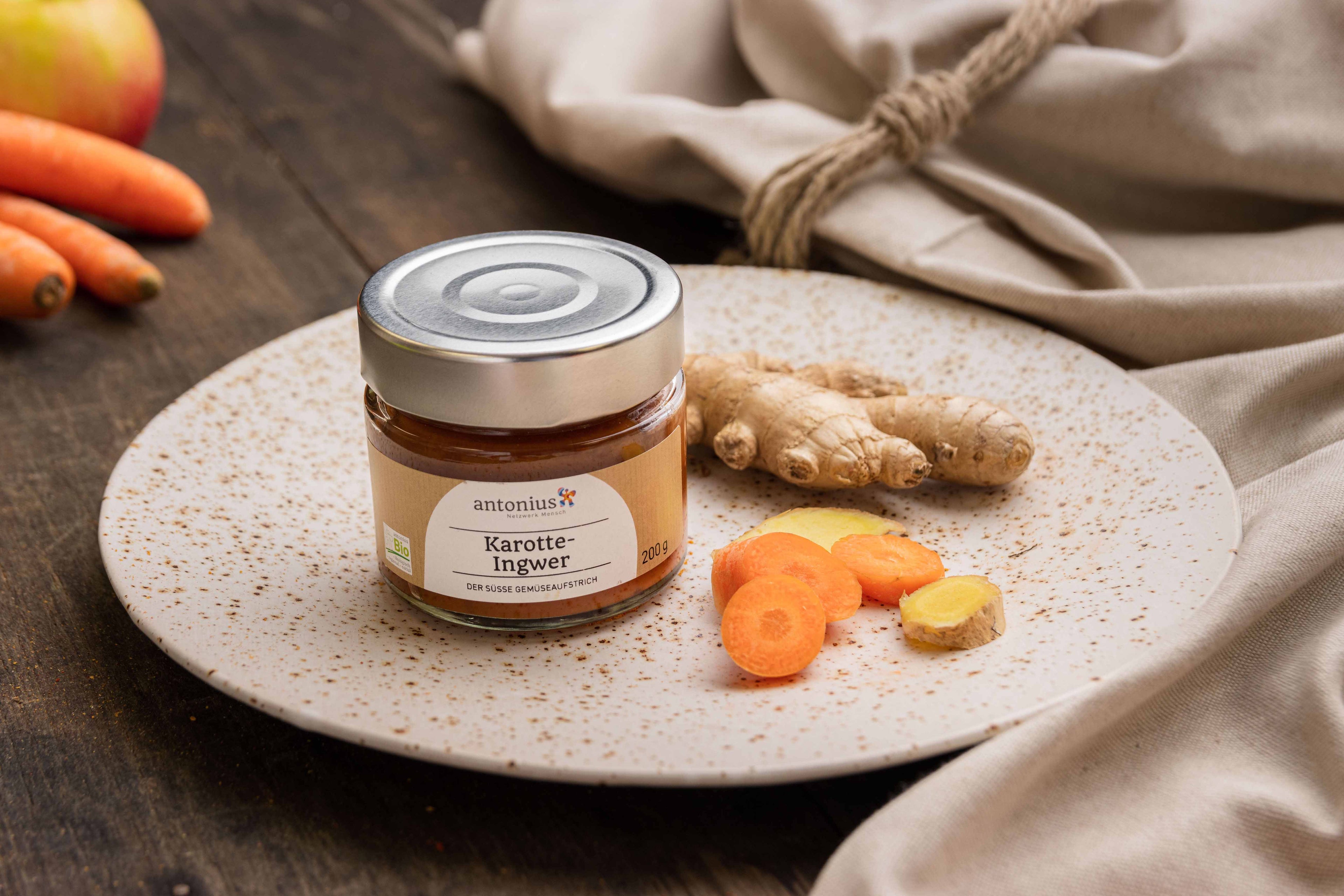 Ein Glas Karotten-Ingwer-Gemüseaufstrich auf einem Teller, angerichtet mit Karotten und Ingwer.