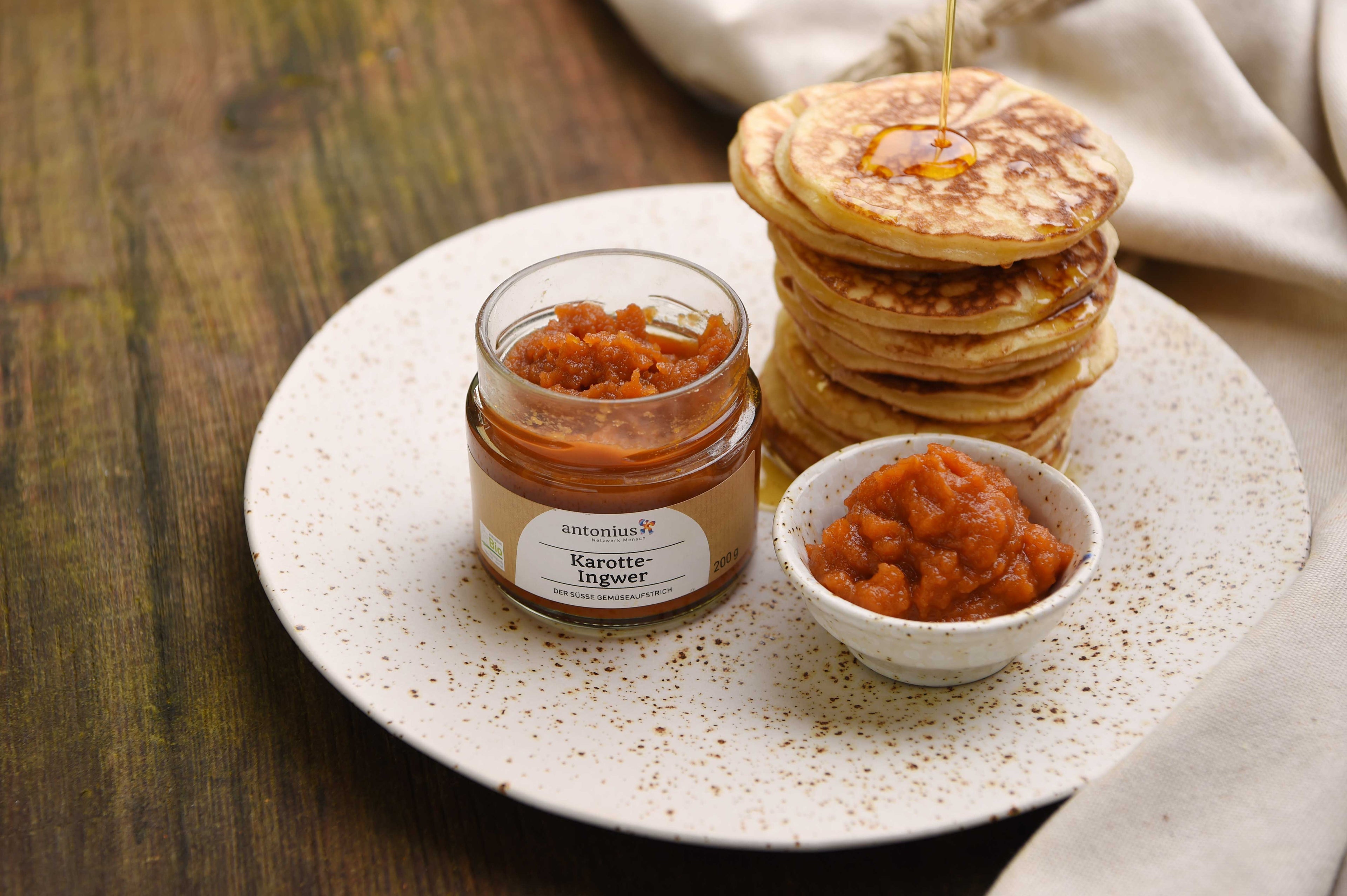 Ein offenes Glas Karotten-Ingwer-Gemüseaufstrich auf einem Teller, angerichtet mit American Pancakes, Ahornsirup und einem Schälchen mit Aufstrich.