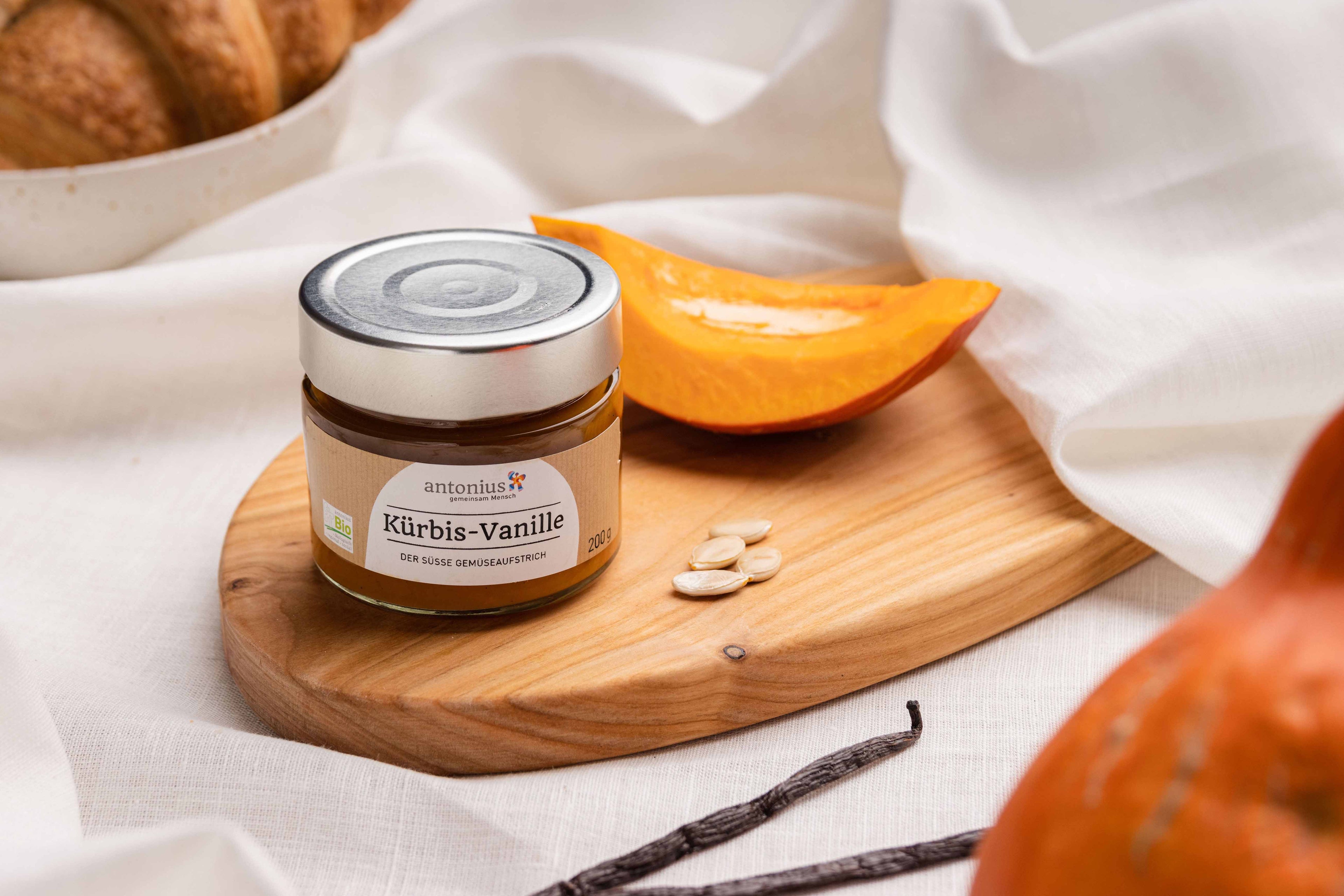 Ein Glas Kürbis-Vanille-Gemüseaufstrich auf einem Frühstücksbrett, angerichtet mit Kürbis, Vanilleschoten und Kürbiskernen.