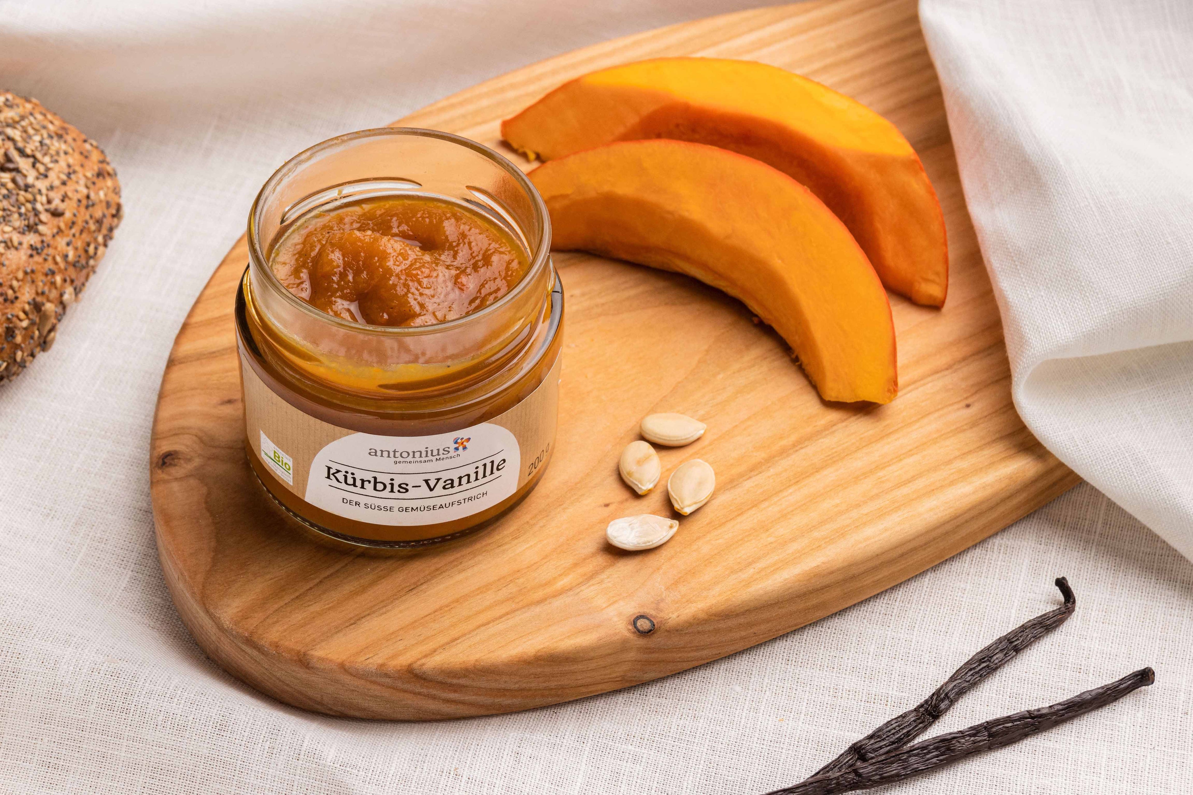 Ein Glas Kürbis-Vanille-Gemüseaufstrich auf einem Frühstücksbrett, angerichtet mit Kürbis, Vanilleschoten und Kürbiskernen. Fotografiert aus der Vogelperspektive.