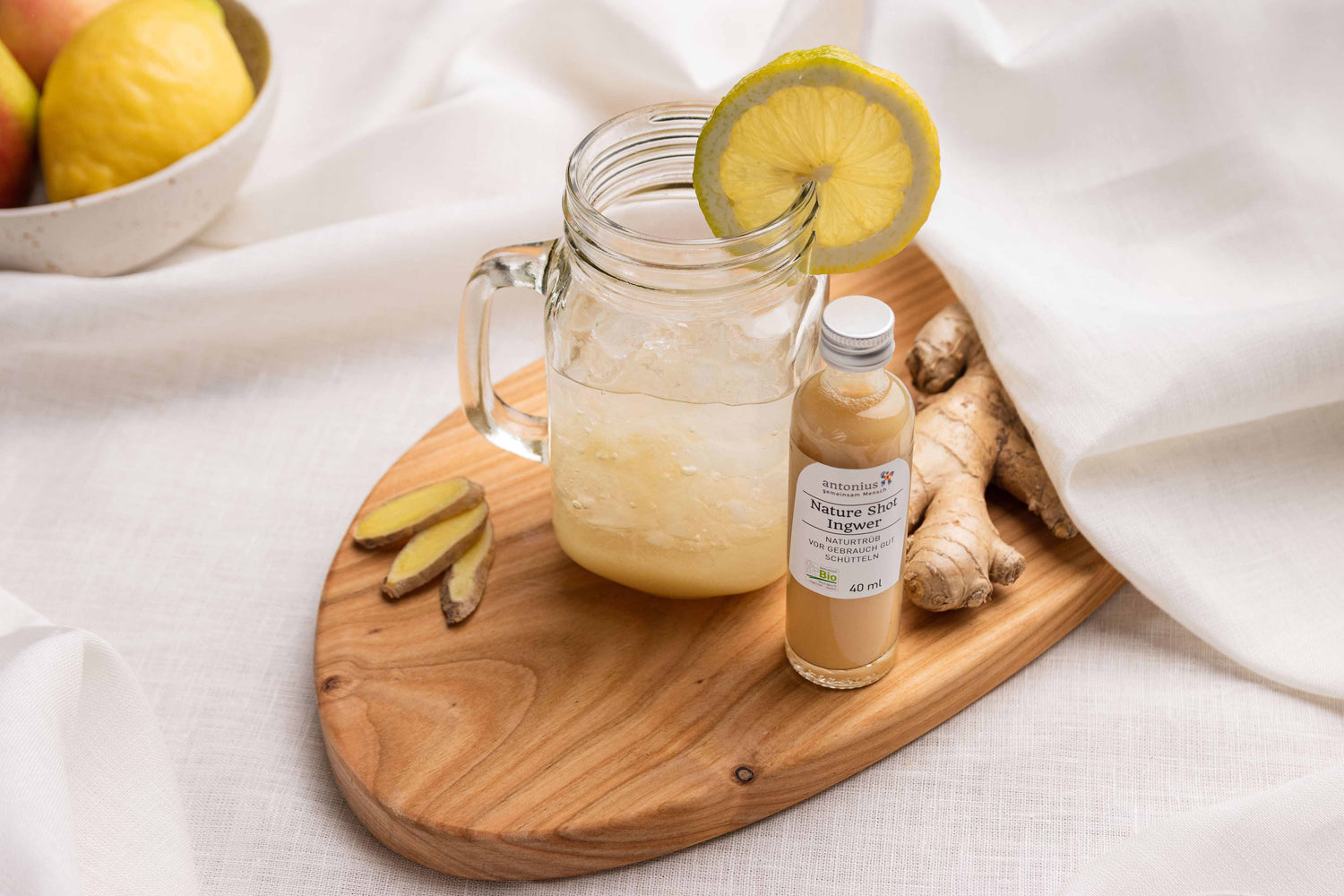Ein Glas mit Henkel ist gefüllt mit Eiswürfeln, Zitronenscheibe und Ingwer Shot. Es steht auf einem Holzbrett neben einer Flasche Inger-Shot und einen Inger-Knolle.