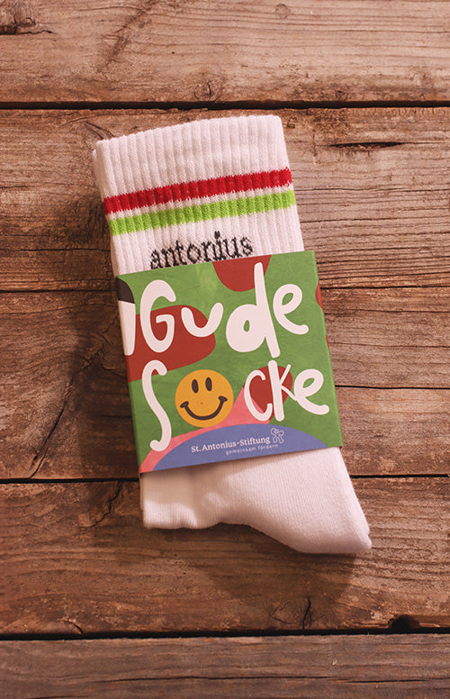 Ein Paar Socken, verpackt mit einem Papierstreifen. Der Papierstreifen liest, Gude Socke, St.Antonius-Stiftung, gemeinsam Mensch. Die socke hat oben einen roten und eine grünen Streifen, zudem die Aufschrift antonius und ein weiterer gelber Streifen, sowie ein dunkler.