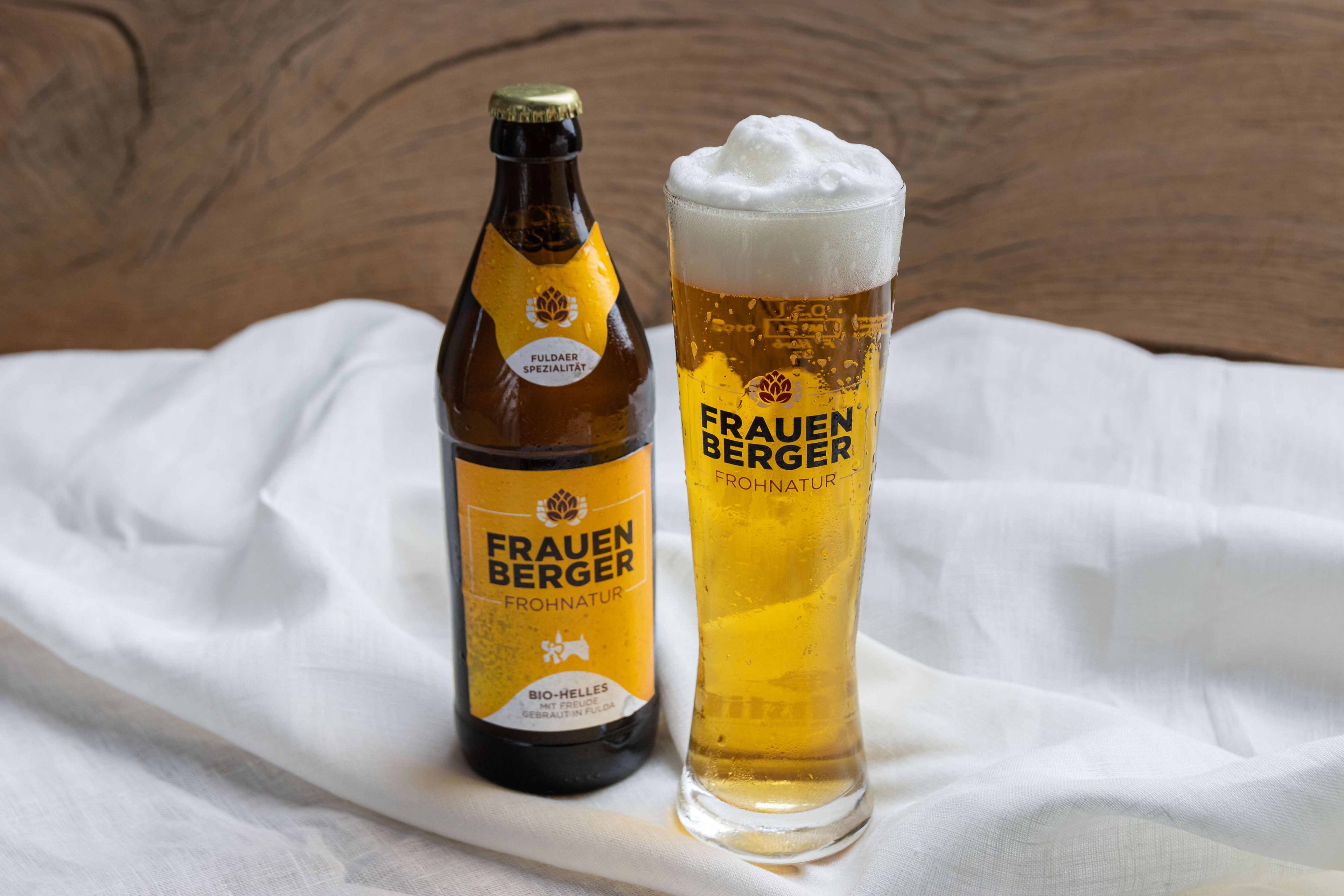 Eine Flasche vom Bio-Bier Frauenberg Frohnatur steht auf einem Tuch. Daneben ein mit Bier und einer Schaumkrone gefülltes Bierglas mit dem "Frauenberger Frohnatur"-Logo.