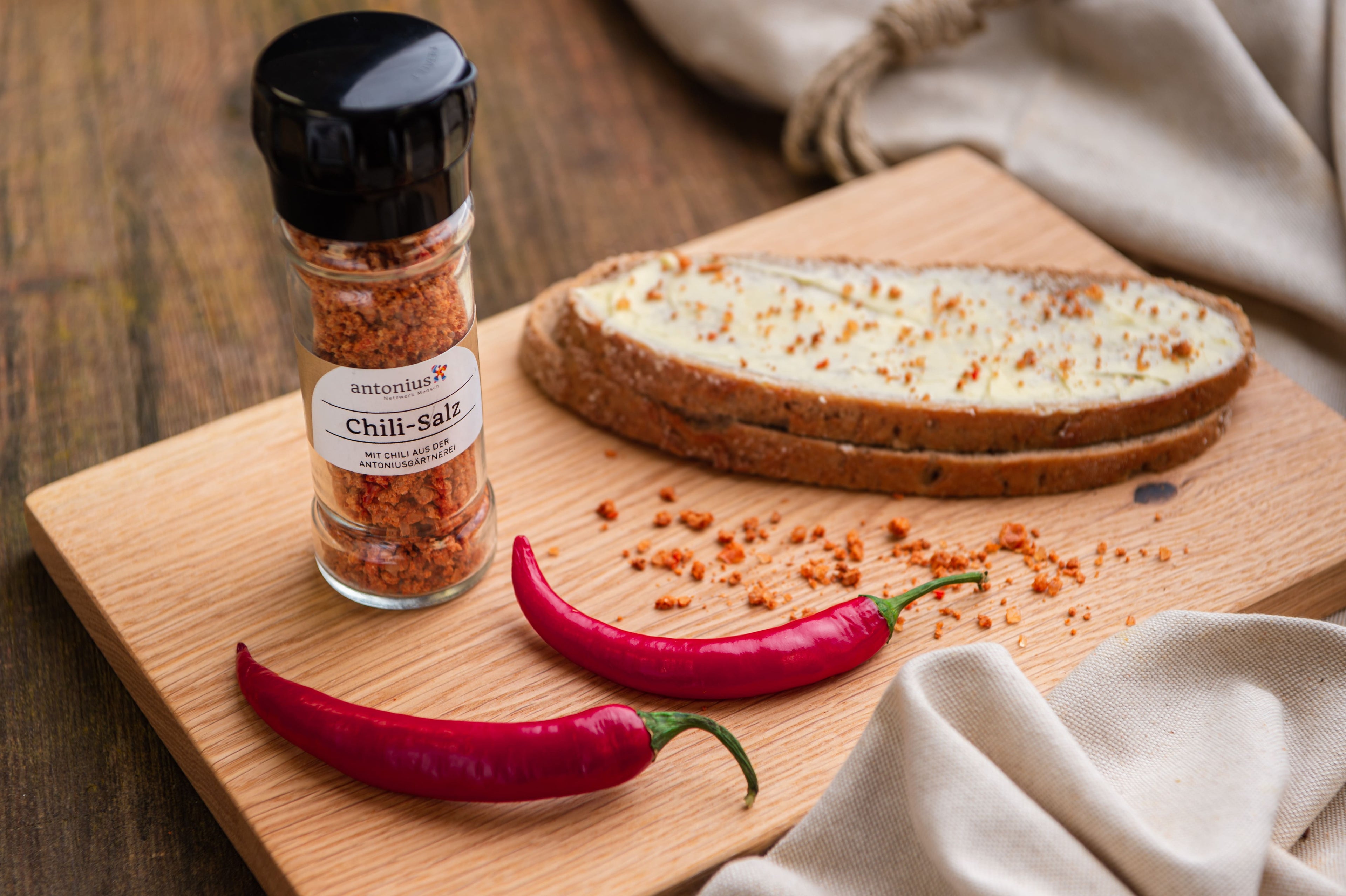 Chili-Salzstreuer auf einem Holzbrett. Daneben ein mit Butter bestrichenes und mit Chili-Salz bestreutes Bauernbrot.
