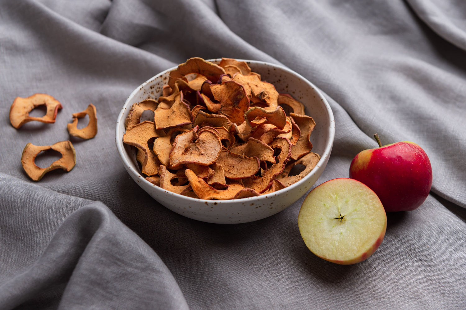 Bio Apfel-Chips aus der Region in einer weiß gesprenkelten Bowl aus Steinzeug-Keramik. Daneben liegt ein aufgeschnittener Apfel zur Dekoration.