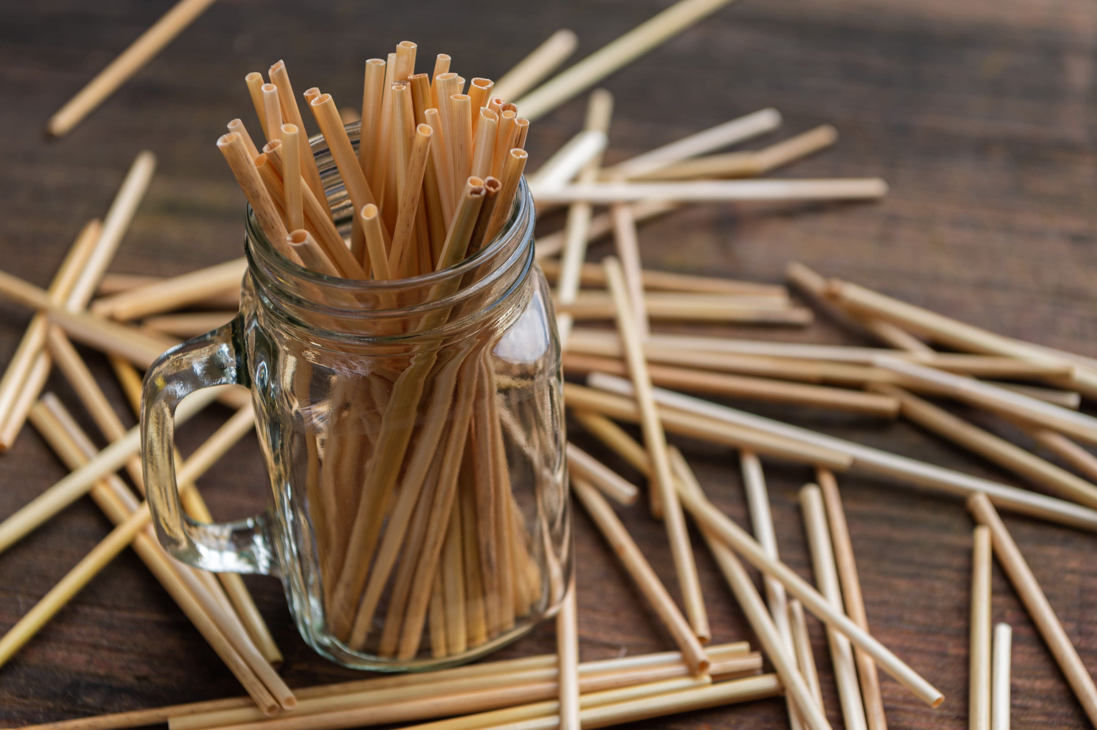 Ein Glas mit Henkel ist mit zahlreichen Strohhalmen aus echtem Stroh gefüllt. Weitere Halme liegen locker auf einem Holztisch verstreut. Die natürliche Farbe und Struktur des Strohs ist gut erkennbar.