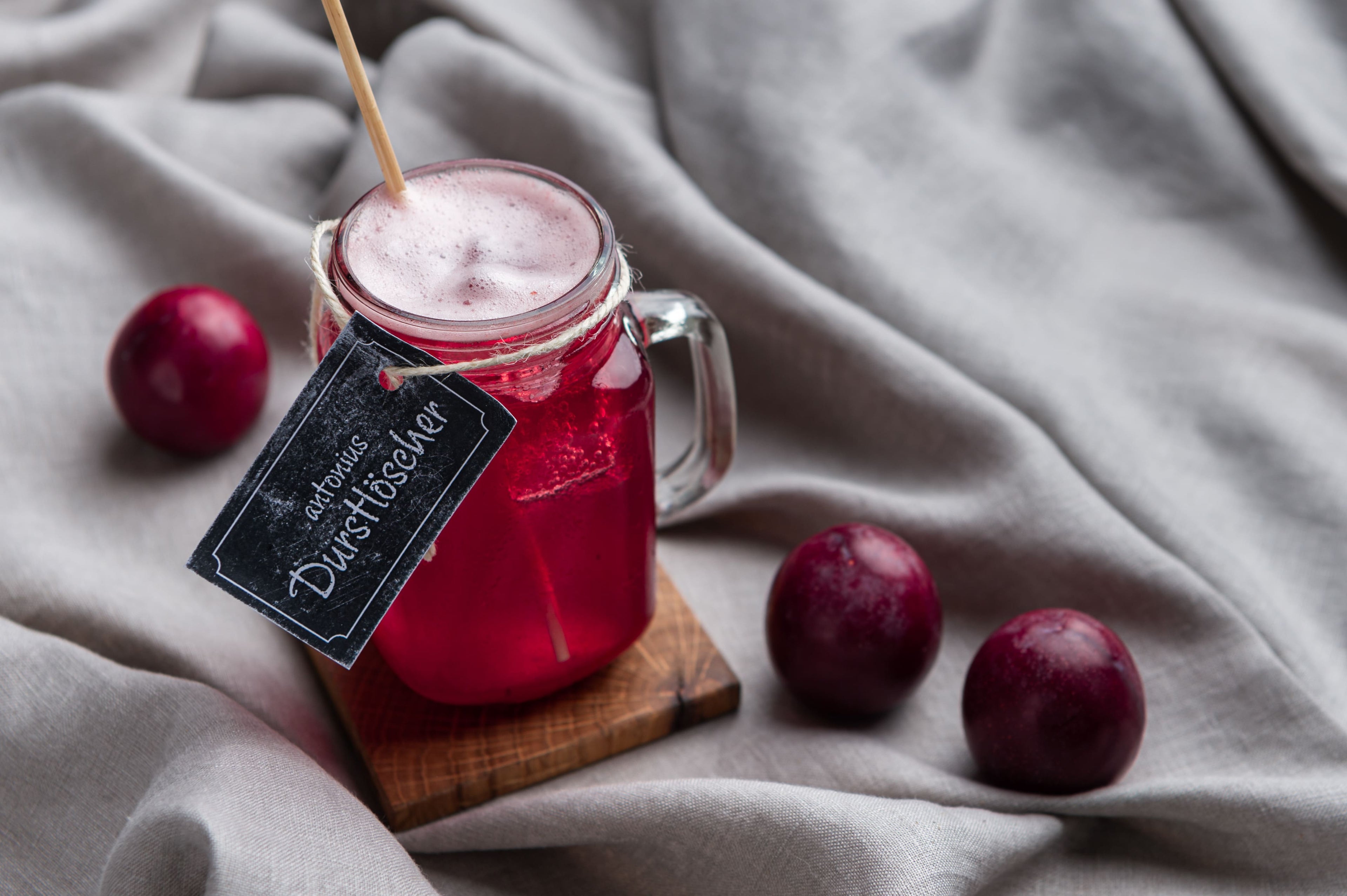 Ein Glas mit Henkel, gefüllt mit einem rot-violetten Getränk und Eiswürfeln, steht auf einem Holzuntersetzer auf grauem Stoff. Im Glas steckt ein Strohhalm aus echtem Stroh. Am Glas hängt ein Schild mit der Aufschrift „antonius Durstlöscher“. Daneben liegen drei dunkelrote Pflaumen.