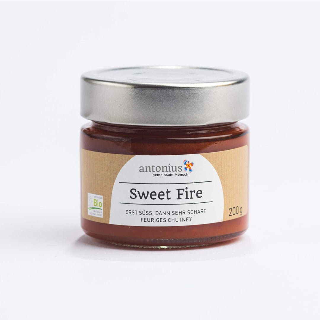 Ein mit einem Metalldeckel verschlossenes Glas mit Sweet Fire-Chutney in Bio-Qualität. Auf dem Etikett sind Informationen zur Füllmenge und die Aufschrift "Sweet Fire - Erst süß, dann sehr scharf - feuriges Chutney"