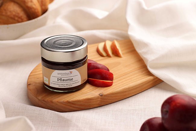 Ein Glas Pflaumen-Fruchtaufstrich auf einem Frühstücksbrettchen, angerichtet mit Pflaumen.