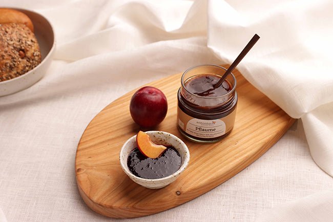 Ein offenes Glas Pflaumen-Fruchtaufstrich auf einem Frühstücksbrettchen, angerichtet mit einem Schälchen mit Aufstrich und Pflaumen.