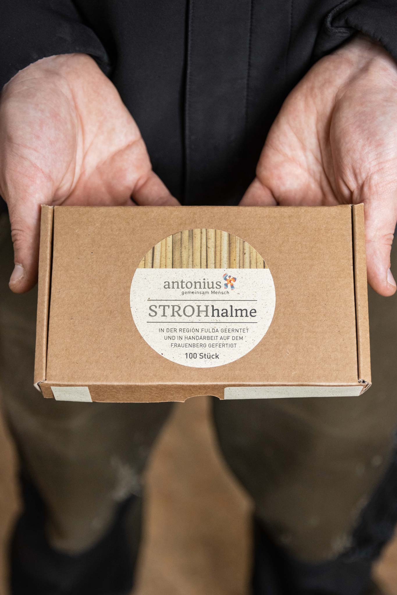 Eine Person hält eine braune Pappschachtel mit Strohhalmen aus echtem Stroh in den Händen. Auf dem Etikett steht: „antonius gemeinsam Mensch, STROHhalme, in der Region Fulda geerntet und in Handarbeit auf dem Frauenberg gefertigt, 100 Stück“.