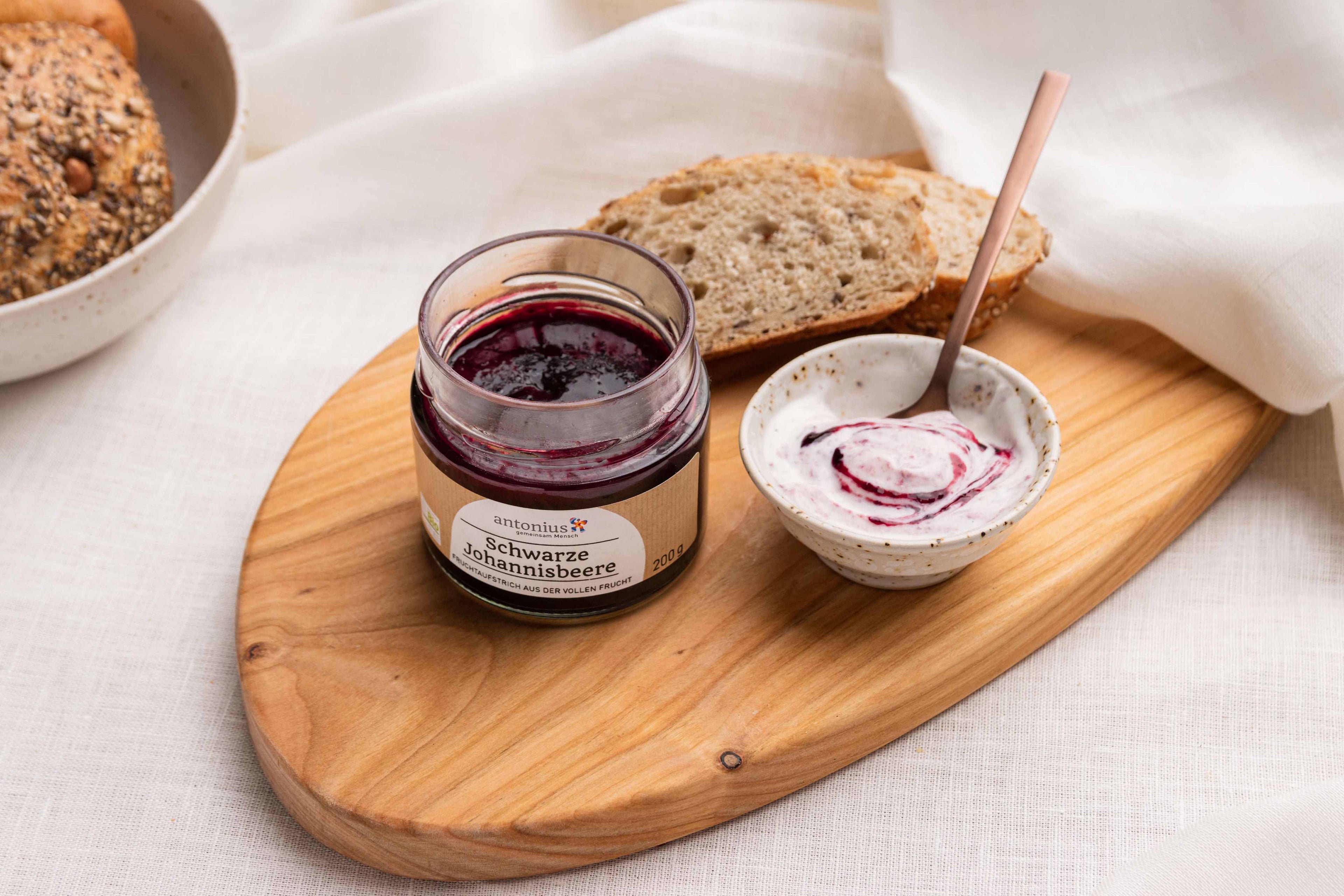 Ein offenes Glas Schwarze Johannisbeere-Fruchtaufstrich auf einem Frühstücksbrettchen, angerichtet mit einer Scheibe Brot und Joghurt.
