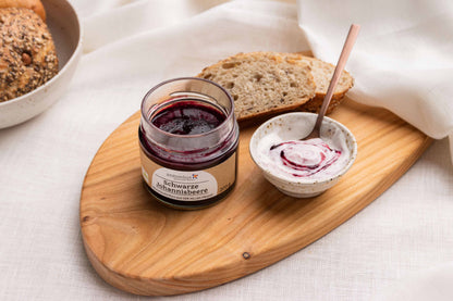 Ein offenes Glas Schwarze Johannisbeere-Fruchtaufstrich auf einem Frühstücksbrettchen, angerichtet mit einer Scheibe Brot und Joghurt.