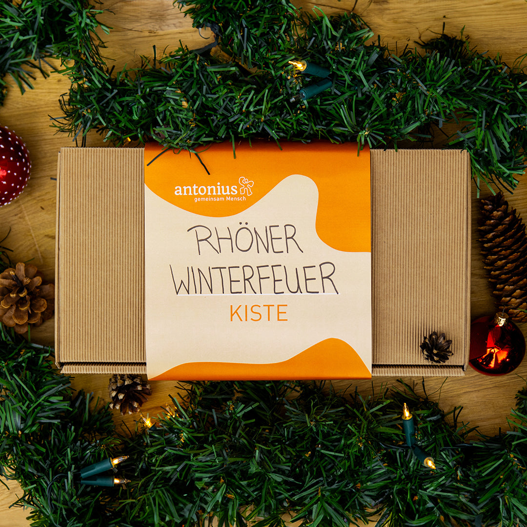 Eine Geschenkkiste aus Pappe und mit einem Streifen aus Papier umbunden. Auf dem Papier kann man antonius, gemeinsam Mensch lesen, sowie Rhöner Winterfeuer Kiste.