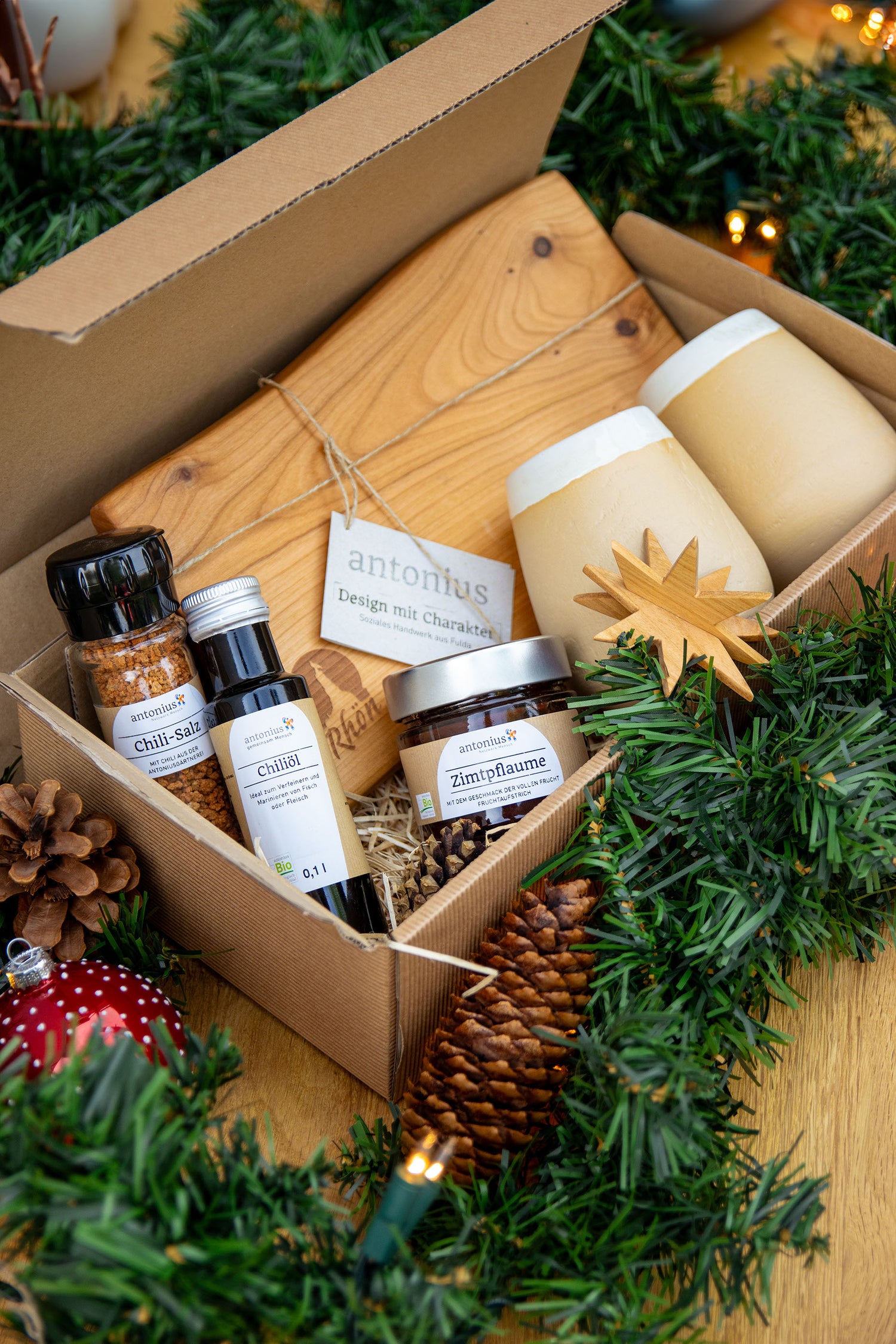 Der Inhalt der Kiste. Gefüllt mit zwei handgemachten Baumassen in Basaltweiß, einem Rhönbrett, einem Stern-Holzanhänger, Chilisalz und Chiliöl und einen Fruchtaufstrich in der Sorte Zimtpflaume. Die Kiste ist gefüttert mit Heu.