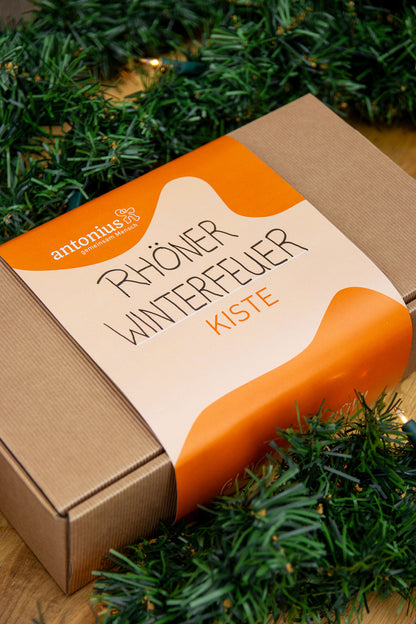 Eine Geschenkkiste aus Pappe und mit einem Streifen aus Papier umbunden Auf dem Papier kann man antonius, gemeinsam Mensch, lesen, sowie Röhner Winterfeuer Kiste.