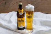 Eine Flasche vom Bio-Bier Frauenberg Frohnatur steht auf einem Tuch. Daneben ein mit Bier und einer Schaumkrone gefülltes Bierglas mit dem "Frauenberger Frohnatur"-Logo. 