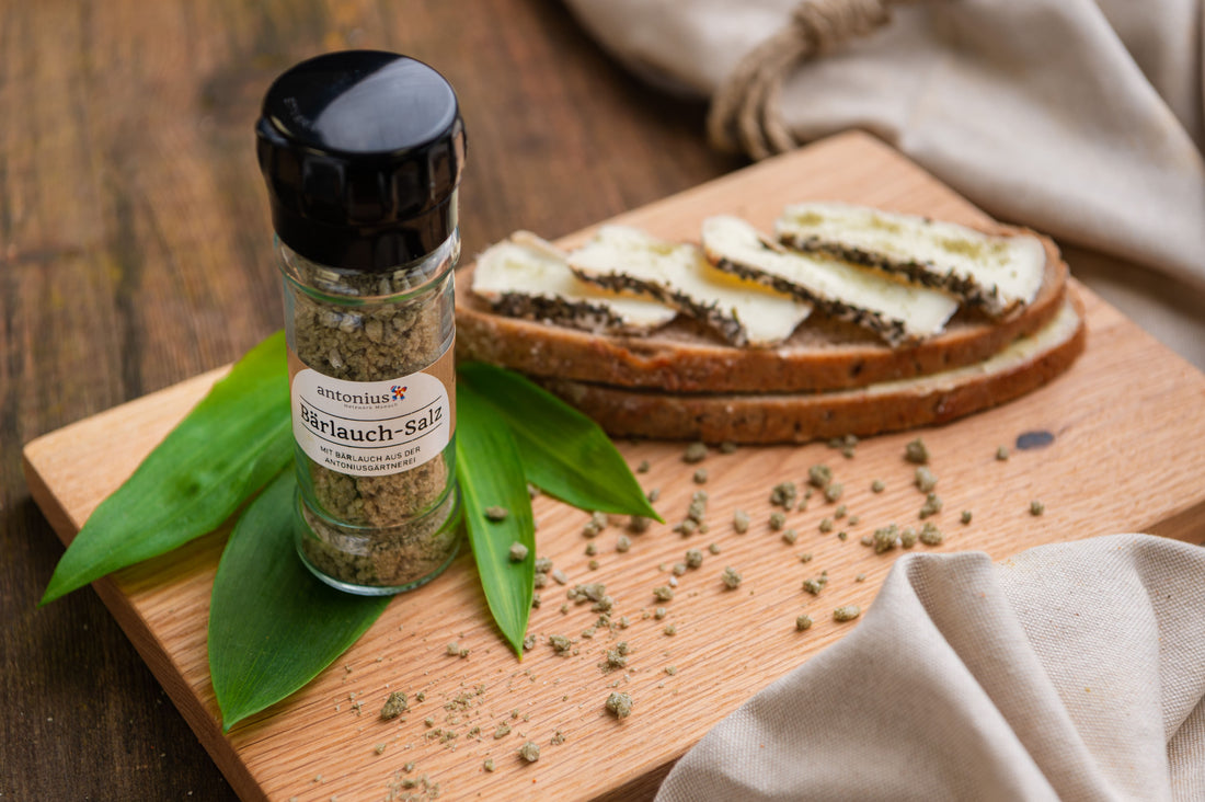Bärlauch-Salzstreuer auf einem Holzbrett. Daneben ein belegtes Bauernbrot mit Käse , bestreut mit Bärlauch-Salz.