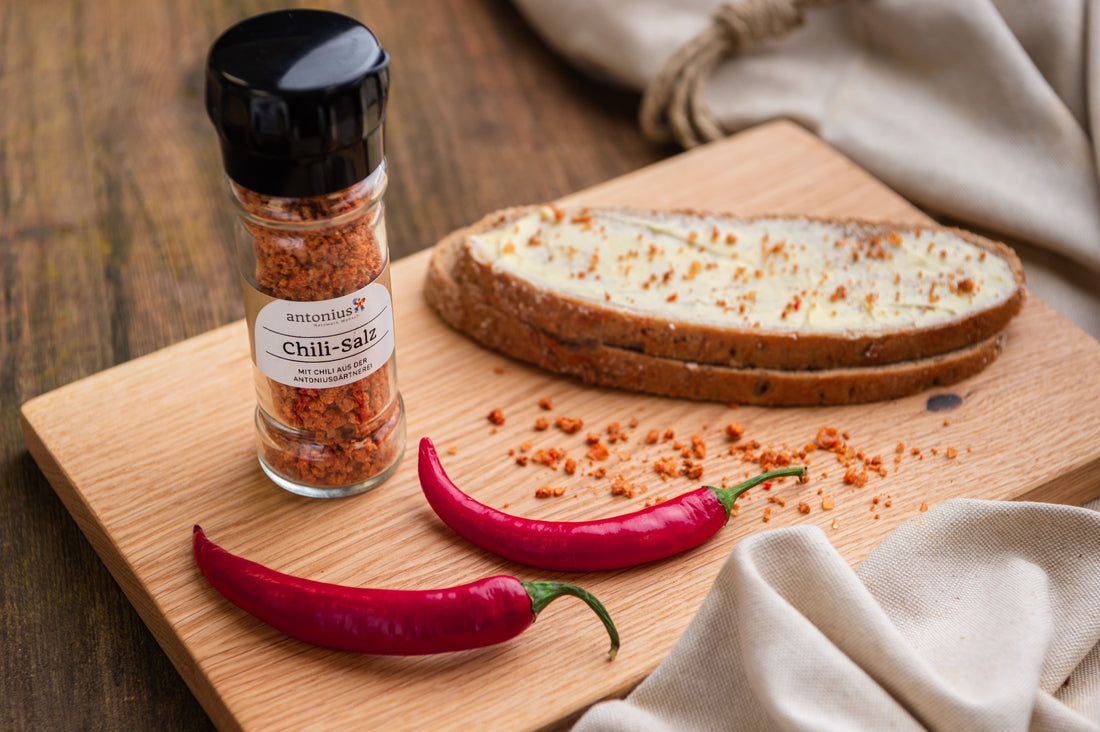 Chili-Salzstreuer auf einem Holzbrett. Daneben ein mit Butter bestrichenes und mit Chili-Salz bestreutes Bauernbrot.